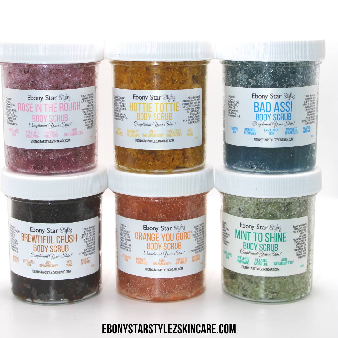 Six jars of body scrubs with different colors and labels indicating various flavors such as Bad Ass, Hottie Tottie, Orange You Gorg', Mint to Shine, and Rose in the Rough.